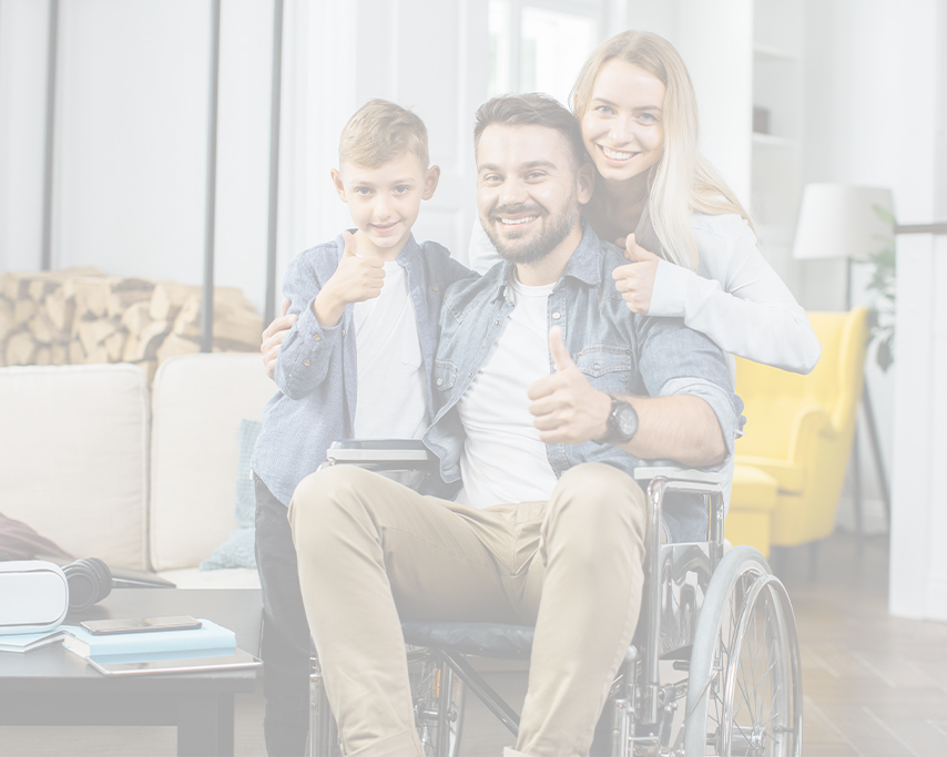 Persona con discapacidad física sentado en una silla de ruedas. Esposa e hijo detrás de él con los dedos pulgares hacia arriba. Tiene una sala en el fondo y están felices.